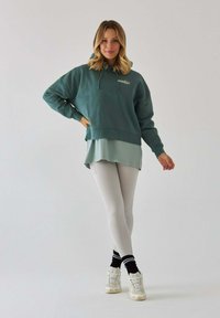 Turkusowa, oversize'owa bluza z kapturem i sznurkiem, noszona na jasnym zielonym topie, zestawiona z białymi legginsami i jasnymi trampkami.