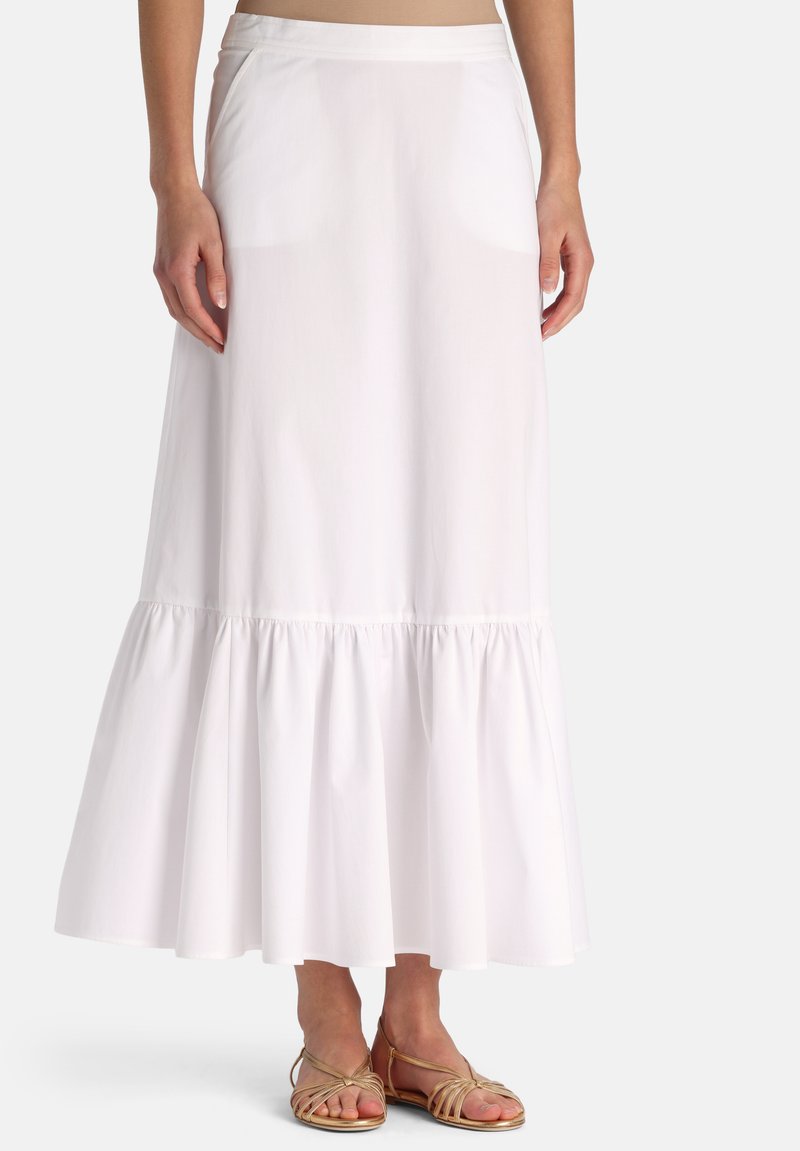 Kvinde iført en lang hvid nederdel med en samlet kant og beige flade sandaler med stropper, stående mod en ensfarvet baggrund.