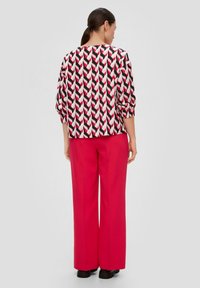 Blouse imprimée en noir, rose et blanc avec un motif en zigzag, à manches trois quarts, associée à un pantalon large rose et des chaussures noires.