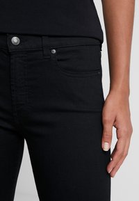 Jeans en denim noir avec une coupe slim, dotés d'une poche avant, de passants de ceinture et d'un bouton métallisé. Le tissu semble résistant avec une texture lisse.