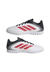 Zapatillas deportivas blancas con acentos negros; presentan tres franjas rojas, parte superior texturizada y una suela moldeada con pequeñas protuberancias de tracción.