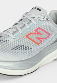 Ανοιχτό γκρι αθλητικό παπούτσι με επάνω μέρος από δίχτυ, λευκά κορδόνια και ροζ προς πορτοκαλί βαθμωτό λογότυπο "N" στο πλάι· κείμενο "1080" στη γλώσσα.