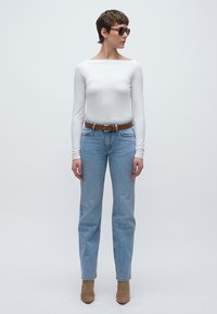 Top alb cu mâneci lungi, blugi cu picior drept, de culoare albastru deschis, curea maro și botine bej. Modelul poartă ochelari de soare închiși la culoare, stând pe un fundal simplu.