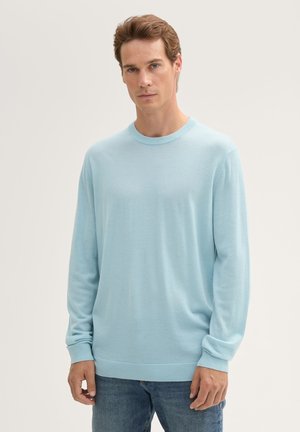 Helle blaue Langarmpullover mit rundem Halsausschnitt sowie gerippten Bündchen und Saum. Der Stoff wirkt weich und hat eine glatte Textur. Über Denim-Jeans getragen.