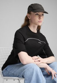 Čierne tričko s krátkym rukávom s šedým prešívaním a logom, skombinované so svetlomodrými džínsami. Na hlave má čiernu čiapku a na ušiach kruhové náušnice.