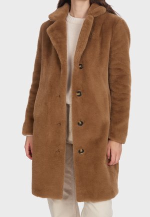 Femme portant un manteau mi-long en fausse fourrure marron avec un large col et quatre boutons foncés sur un pull beige clair et un pantalon.