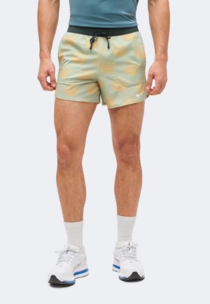 Άνδρας φορώντας πράσινα και πορτοκαλί μοτίβο σορτς για τρέξιμο Nike με μαύρο κορδόνι, λευκές κάλτσες και λευκά αθλητικά παπούτσια σε μονόχρωμο φόντο.