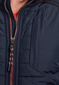 Muž ve tmavě modré prošívané bundě s černým a červeným zipem a červenou texturovanou vnitřní vrstvou viditelnou u límce.