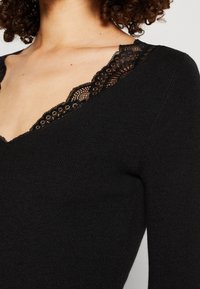 Gros plan sur un haut noir à manches longues avec un bord en dentelle le long du décolleté en V, porté par une personne aux cheveux bouclés.