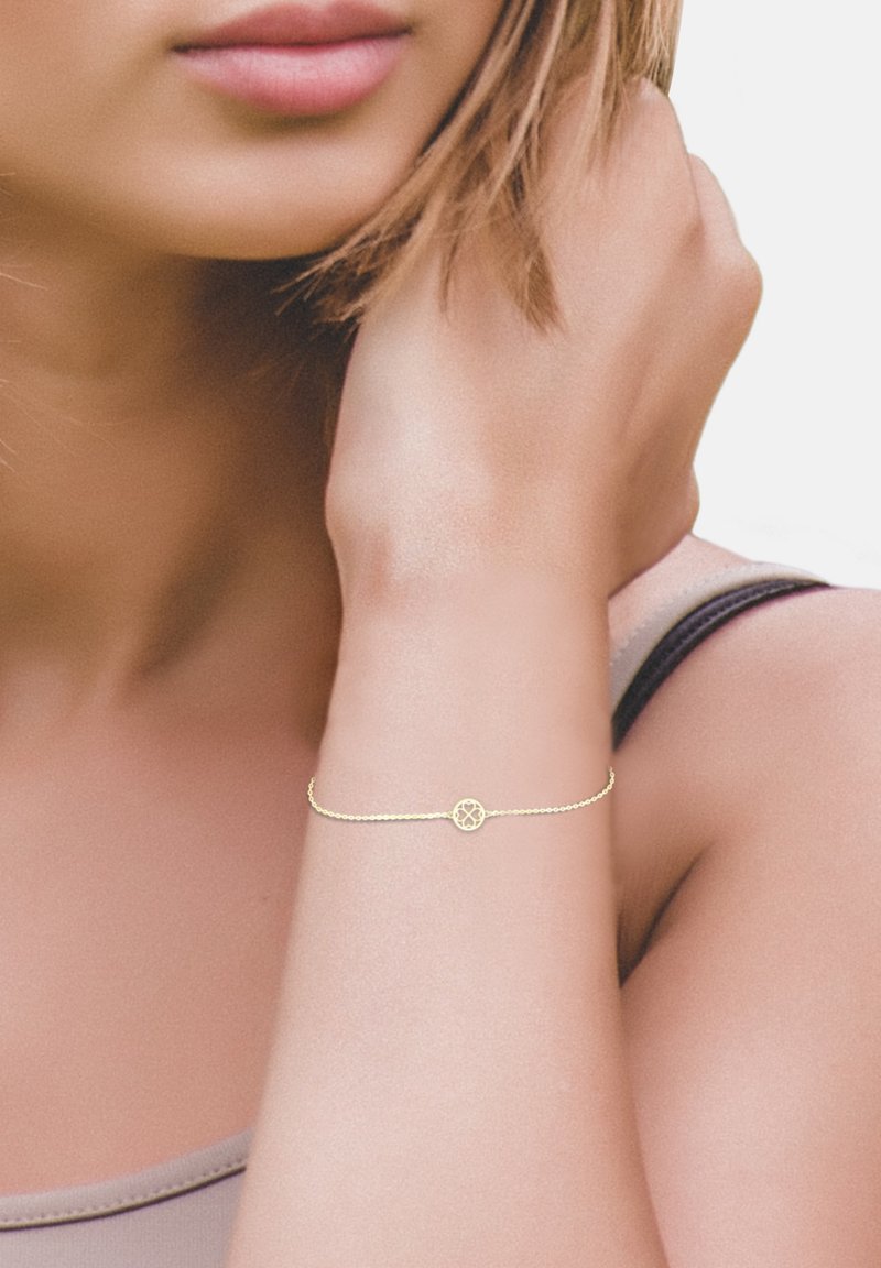 Bracciale in catena d'oro con un pendente floreale circolare, indossato su un polso di pelle chiara, con una texture liscia e una leggera lucentezza.