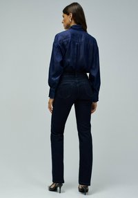 Camisa de mezclilla azul marino con mangas largas, jeans oscuros ajustados y tacones negros, vista desde la parte trasera contra un fondo gris claro.