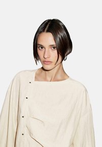 Blouse crème ample avec un décolleté carré, ornée de boutons et d'une texture subtile. Le tissu semble léger et doux.