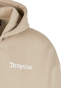 Beige hættejakke lavet af blødt stof med et stort, broderet "Dropsize" logo i hvid, med snoretræk i hætten og en afslappet pasform.