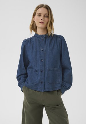 Femme portant une veste en jean bleue boutonnée avec deux poches avant et un pantalon vert olive, debout devant un fond clair uni.
