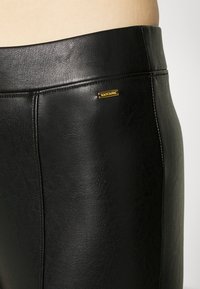 Mustat keinonahkapantsit, joissa on sileä pinta, näkyvä sauma ja kultasävyinen logolevy vyötäröniemessä.