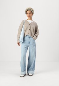 Hellbrauner Strickcardigan mit drei Knöpfen, kombiniert mit hellblauen weit geschnittenen Jeans. Accessoires mit einer hellblauen Schultertasche. Weiße Sneakers.