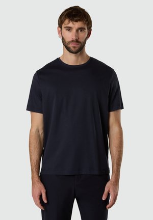 Uomo con capelli castani corti e barba, che indossa una maglietta nera a maniche corte semplice e pantaloni neri, in piedi davanti a uno sfondo grigio chiaro.