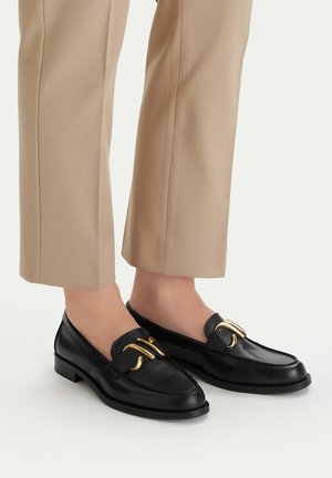 Czarne skórzane loafersy z ozdobnymi złotymi dodatkami, noszone z beżowymi spodenkami o długości 7/8 na bose stopy.