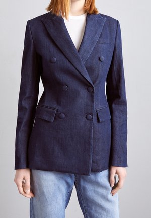 Person trägt einen dunkelblauen, zweireihigen Blazer mit stoffbezogenen Knöpfen über einem weißen Oberteil und hellblauen Jeans.