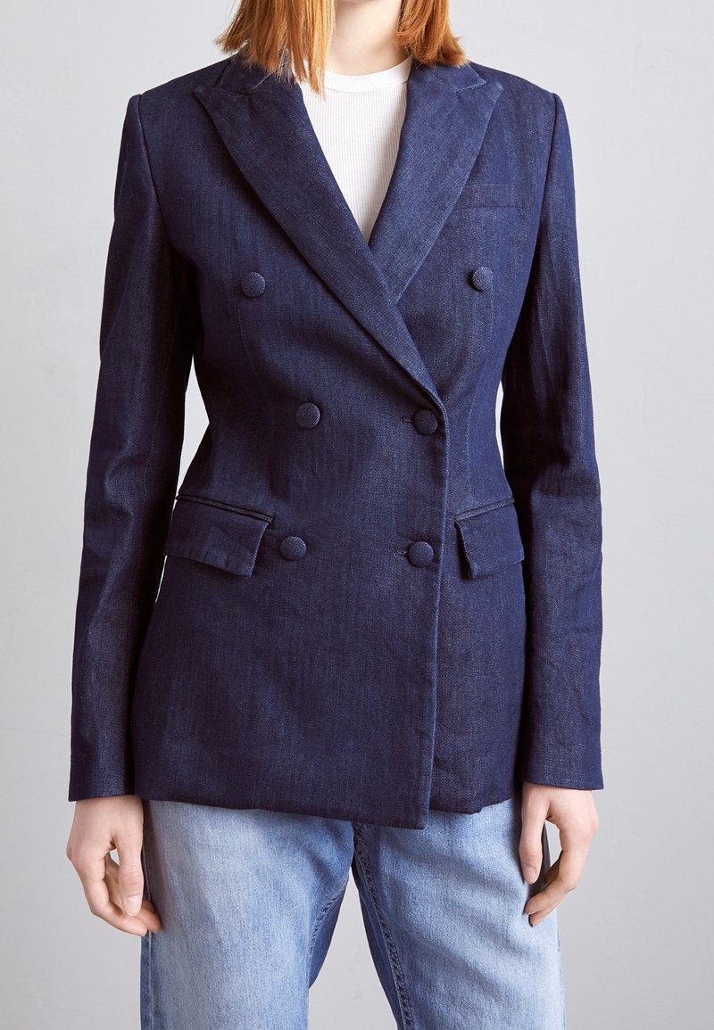 Personne portant un blazer croisé bleu foncé avec des boutons recouverts de tissu, sur un haut blanc et un jean bleu clair.