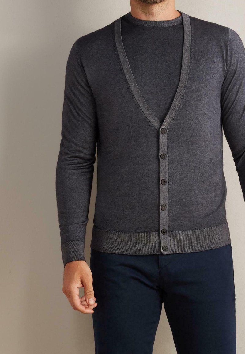 Homme portant un cardigan boutonné col V gris foncé par-dessus une chemise à manches longues assortie et un pantalon bleu marine, debout devant un fond neutre.