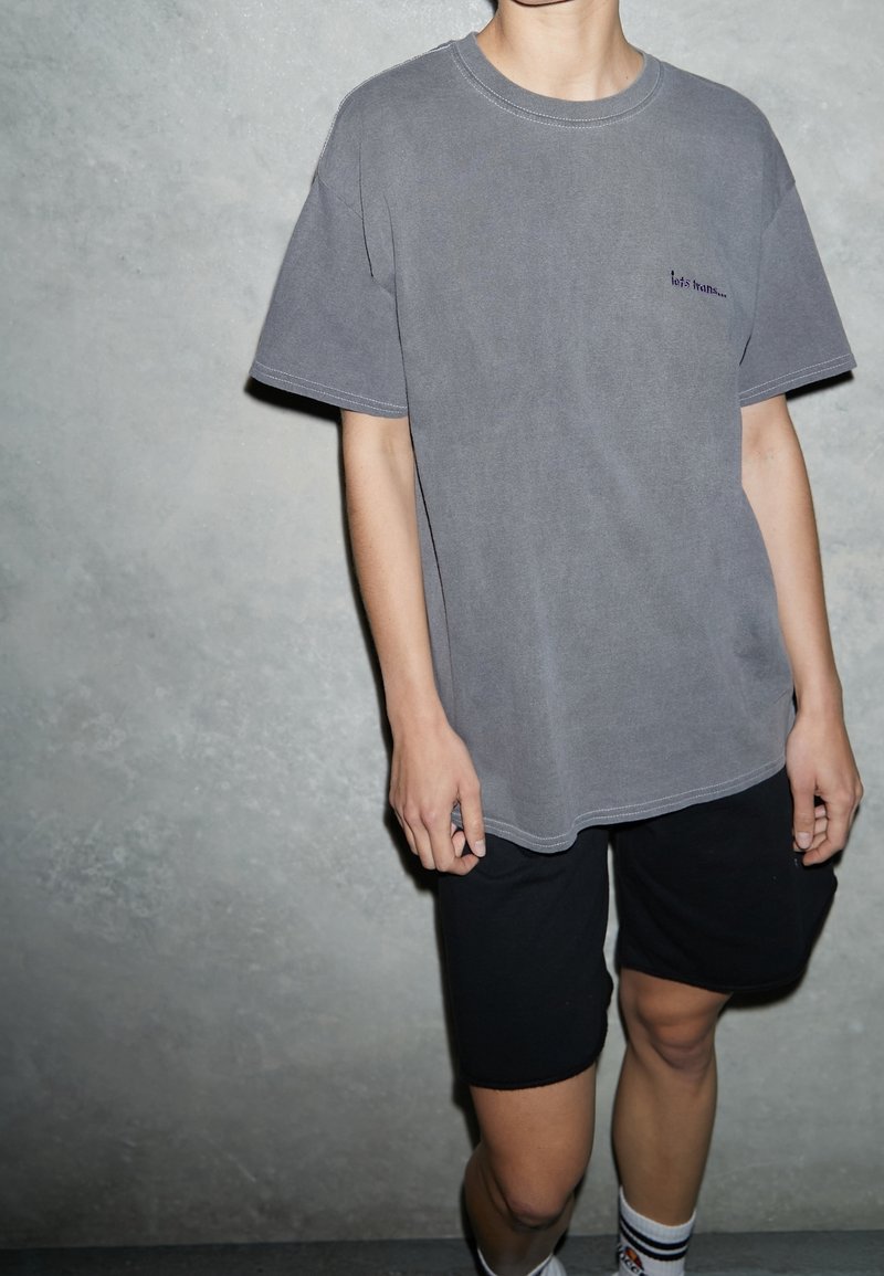 Personne portant un t-shirt gris ample avec un petit texte et un short noir, debout contre un mur gris texturé.