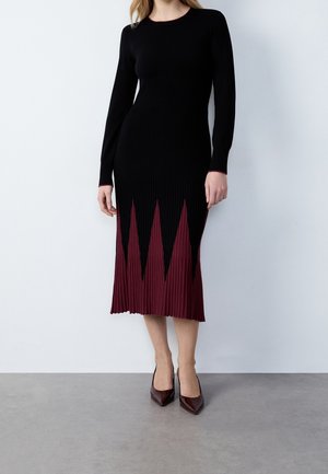 Femme portant une robe midi noire à manches longues avec un ourlet plissé triangulaire bordeaux et des escarpins pointus bordeaux, debout sur un sol gris.