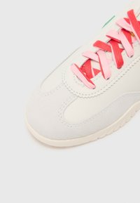 Gros plan sur l’avant d’une basket blanche avec des lacets croisés rouges et roses et des détails de couture légers sur un fond clair.