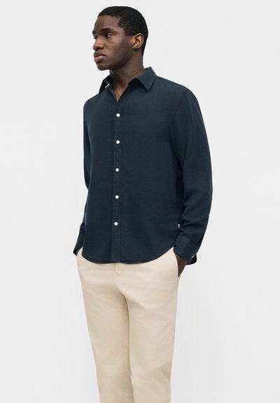 Άνδρας που στέκεται με τα χέρια στις τσέπες, φορώντας σκούρο μπλε πουκάμισο με κουμπιά και μπεζ παντελόνι, μπροστά σε απλό φόντο.