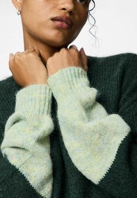 Maglione verde lavorato a maglia con un corpo testurizzato e maniche a costine verde chiaro con un motivo a macchie. Mani incrociate all'altezza del collo.