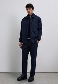 Camisa de botón azul marino con dos bolsillos en el pecho, combinada con pantalones a juego. Zapatillas negras y camiseta interior gris claro visible en el cuello.