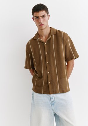 Mand iført brun kortærmet skjorte med lodrette hvide striber og lyseblå jeans, stående mod en hvid baggrund.