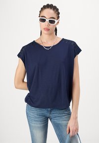 Marineblå t-skjorte med korte ermer i teksturert stoff, blondeakenter på skuldrene, matchet med lyseblå jeans og en sølvkjede.