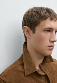 Braune Wildlederjacke mit spitzem Kragen, Druckknöpfen und weicher Textur. Das Profil des Models ist vor einem neutralen Hintergrund zu sehen.
