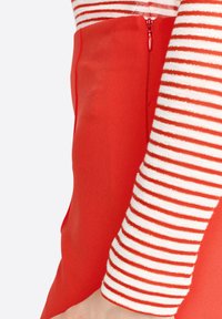 Punat punah merah dengan ritsleting samping, dipasangkan dengan atasan lengan panjang yang memiliki garis horizontal merah dan putih. Tekstur kain yang halus terlihat.