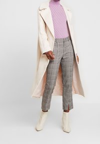 Un long manteau beige clair sur un pull à col roulé lilas, assorti à un pantalon à carreaux gris avec des fentes. Des bottines blanches complètent la tenue.