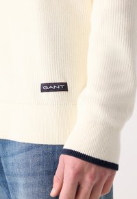 Sweter w kolorze kremowym, ryżowym, z granatowymi wykończeniami na mankietach oraz naszywką z logo GANT w dolnym lewym rogu. Noszony na jasnoniebieskich dżinsach.