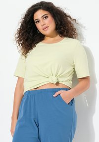 Svetle zelený uzlový crop top s krátkým rukávem, zkombinovaný s volnými modrými kalhotami s bočními kapsami. Měkká, hladká textura látky.