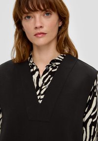 Top senza maniche nero con scollo a V profondo sopra una camicia con bottoni caratterizzata da un motivo a strisce bianche e nere a zebra. Tessuto liscio.