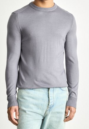 Homme portant un pull ajusté à manches longues gris clair avec des poignets côtelés et un jean bleu clair, debout devant un fond blanc uni.