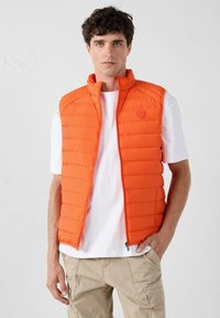 Oranje gewatteerde vest met verticale gestikte patroons, hoge kraag en ritssluiting aan de voorkant. Gedragen over een wit T-shirt, gecombineerd met lichte cargoshorts.