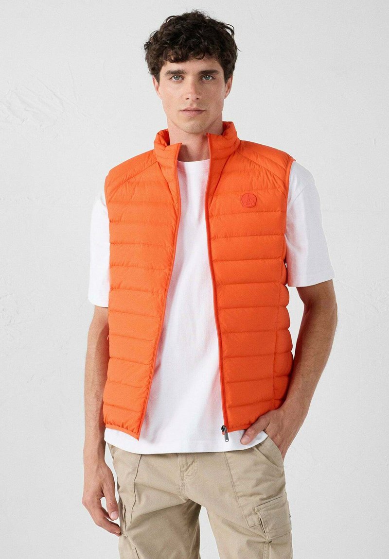 Oranje gewatteerde vest met verticale gestikte patroons, hoge kraag en ritssluiting aan de voorkant. Gedragen over een wit T-shirt, gecombineerd met lichte cargoshorts.