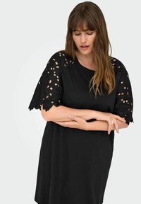 Woman wearing black dress with lace cutout sleeves, arms crossed, looking downward against a plain light background.