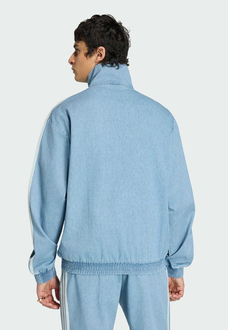 Jonge man met donker krullend haar, gekleed in een lichtblauw denim trainingspak met elastische manchetten en witte zijstrepen, gezien van achteren.