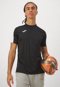 Camisa de deporte negra con mangas cortas blancas, que presenta un logo en el pecho. La persona sostiene un balón de fútbol vibrante en naranja y negro.