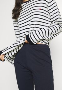 Cămașă cu mânecă lungă, dungi alb-negru, cu un mic logo roșu; asociată cu pantaloni ajustați navy blue, cu un material moale și linii curate.