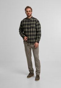 Karierter Flanellshirt in Dunkelgrün und Schwarz mit einer Brusttasche, kombiniert mit beigen Hosen und braunen Schuhen. Einfaches, strukturiertes Design.