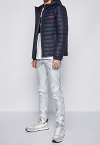 Marineblå pufferjakke med quiltet tekstur, grå metallic jeans og grå-hvide sneakers med sorte detaljer. Minimalistisk design, afslappet pasform.