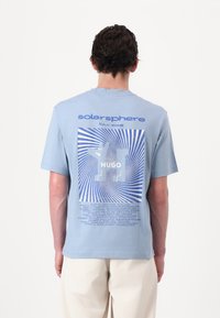 Lyseblå bomulls t-skjorte med grafisk design med konsentriske linjer, tekst "solarsphere tour 1998," og et stort logo på baksiden.