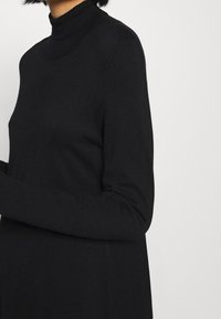 Femme portant un pull noir ajusté à col roulé à manches longues, sur un fond clair uni, vue des épaules aux hanches.